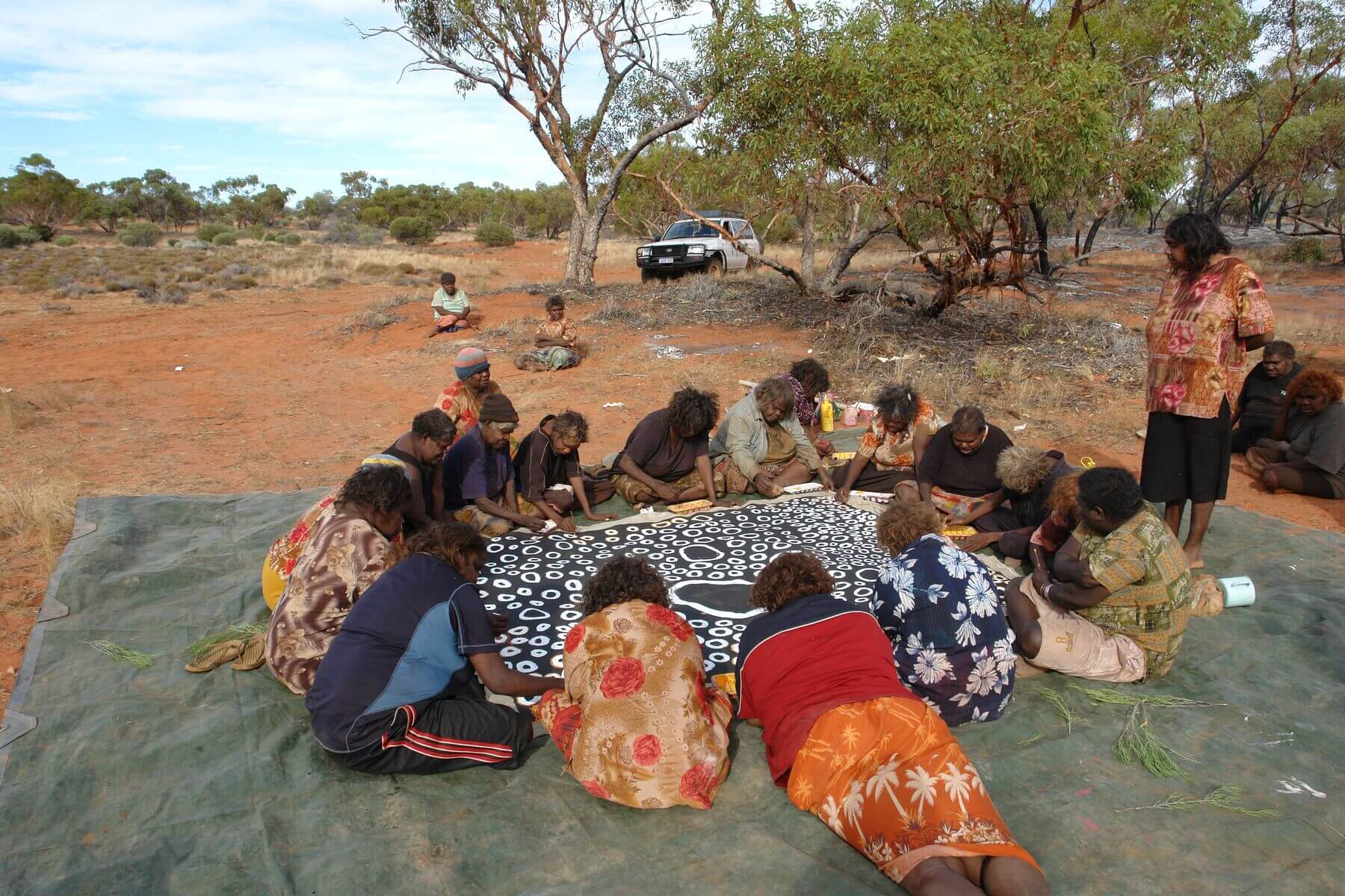 18 women from Spinifex Arts sitting and painting on a large black and white canvas on the ground.